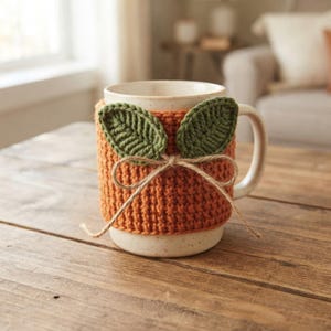 Peut inclure: Une tasse en céramique couleur crème dotée d'une housse en crochet orange texturée. La housse est ornée de deux feuilles crochetées vert foncé et d'une fine ficelle marron nouée en nœud au centre, s'ajustant parfaitement autour de la tasse.