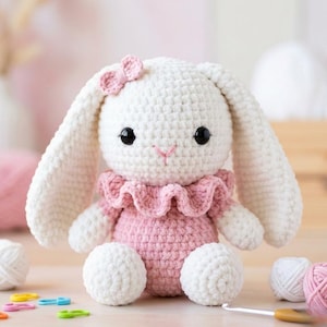 May include: A handmade crochet bunny doll in white and pink. The bunny has long, floppy ears, black eyes, and a pink bow. It wears a pink dress with a ruffled collar. The bunny is sitting with its arms and legs extended.