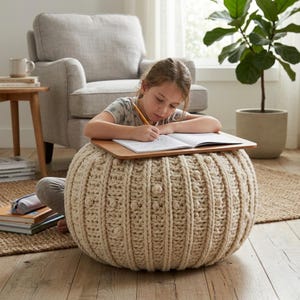 Może przedstawiać: Kremowy, dziergany puf z drewnianym stolikiem na laptopa. Okrągły puf ma teksturowany, żebrowany wzór. Osoba siedzi na podłodze, używając pufa jako biurka do pisania w notatniku ołówkiem. Scena rozgrywa się w salonie.