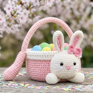 Può includere: Un cestino pasquale all'uncinetto rosa e bianco, con un coniglietto attaccato. Il cestino è pieno di uova di Pasqua colorate. Il coniglietto ha orecchie foderate di rosa e un fiocco rosa. Anche il manico del cestino è rosa.