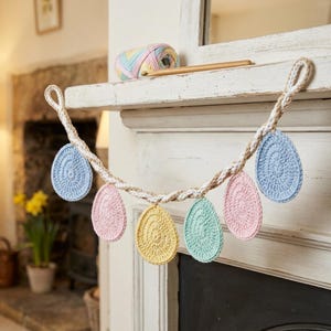 Puede incluir: Una guirnalda de Pascua hecha a mano con seis adornos de huevo de ganchillo en colores pastel: azul, rosa, amarillo y verde. Los huevos están suspendidos de una cuerda trenzada. Una madeja de hilo y agujas de tejer descansan en una repisa.