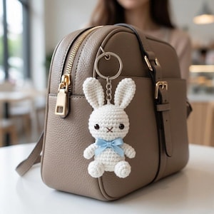May include: A white crochet bunny keyring with a blue bow is attached to a taupe leather handbag. The bunny has black eyes and a stitched mouth. The handbag has a gold zip and hardware.