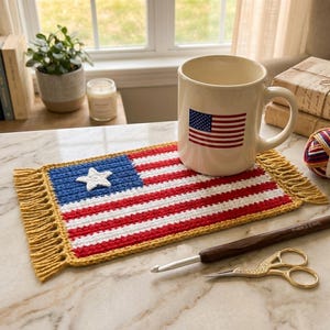 May include: A cream-colored mug with an American flag design sits on a crocheted mat resembling the American flag. A crochet hook and gold scissors are next to the mat. The mat has a blue square with a white star and red and white stripes.