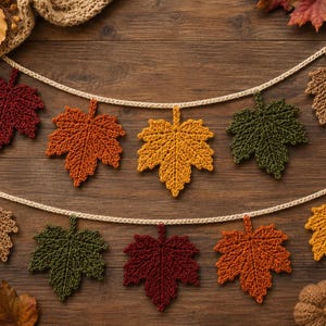 May include: A decorative garland featuring two rows of crocheted maple leaves in autumn colors including deep red, burnt orange, golden yellow, forest green, and tan. The leaves are attached to a cream-colored crocheted string for hanging.