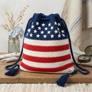 Puede incluir: Bolso de ganchillo hecho a mano con los colores de la bandera estadounidense. El bolso tiene una parte superior azul con estrellas blancas, rayas rojas y blancas, y cordones azul marino con borlas. El bolso está hecho de hilo.