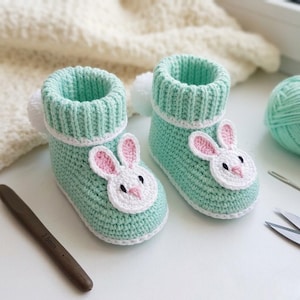 May include: A pair of handmade, mint green crochet baby booties. Each bootie features a white and pink bunny face applique on the toe, a white ribbed cuff, and a small white pom-pom on the back. A crochet hook and yarn are visible.