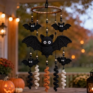 May include: A hanging mobile featuring five crocheted black bats of varying sizes suspended from a circular wooden hoop. The bats have white button eyes and are accented with black and beige beads, with dangling spiral tails in black, orange, and cream.