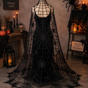 Könnte beinhalten: Ein schwarzer Häkelumhang mit Spinnennetz-Muster und integrierten Spinnenmotiven. Der Umhang liegt über einem schwarzen Kleid auf einer Schaufensterpuppe und reicht bis zum Boden, mit einem breiten, gebogten Saum und einem hohen Kragen.