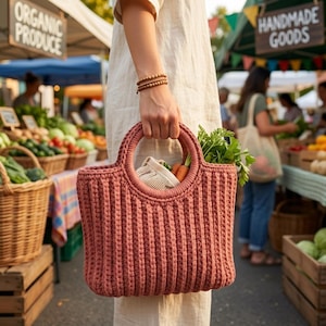 May include: A dusty rose crochet tote bag with a ribbed texture and two circular handles. The bag contains a small mesh produce bag and fresh green leafy vegetables with orange carrots visible inside the main compartment.