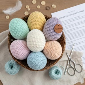 May include: A wicker basket holds seven pastel-coloured, crocheted Easter eggs. The eggs are in shades of blue, pink, yellow, and white. One egg has a "Handmade" tag. The image also shows yarn, scissors, and a pattern sheet for Easter eggs.