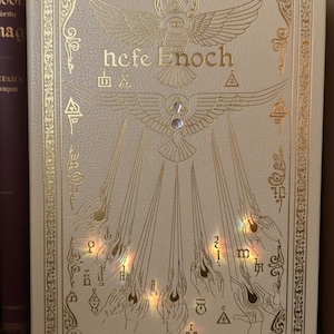 May include: Cream-colored book cover with gold foil accents. The title "hefe Enoch" is displayed above an illustration of a bird with outstretched wings. The cover features ornate borders and symbols.