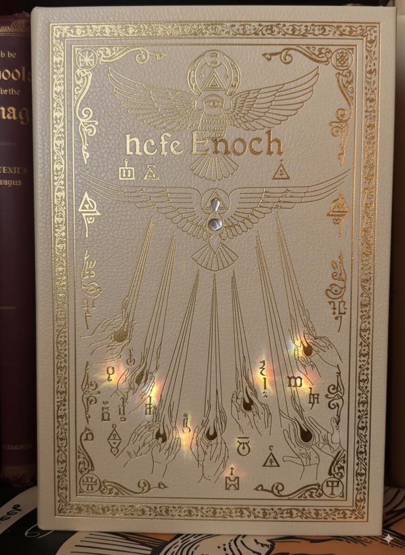 May include: Cream-colored book cover with gold foil accents. The title "hefe Enoch" is displayed above an illustration of a bird with outstretched wings. The cover features ornate borders and symbols.