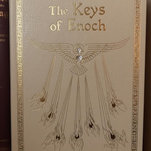 May include: Cream-colored book titled "The Keys of Enoch" with gold lettering and decorative border. The cover features a gold emblem of a bird with outstretched wings, a triangle, and rays emanating from hands.