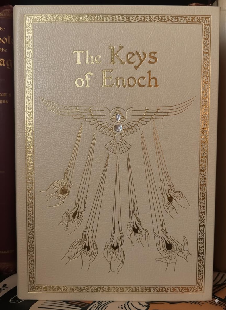 May include: Cream-colored book titled "The Keys of Enoch" with gold lettering and decorative border. The cover features a gold emblem of a bird with outstretched wings, a triangle, and rays emanating from hands.