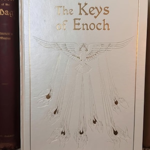 Könnte beinhalten: Ein weißes Hardcover-Buch mit dem Titel "The Keys of Enoch" mit goldener Schrift und geprägten Illustrationen. Der Einband hat einen goldenen Rand und zeigt eine Taube mit ausgebreiteten Flügeln und Strahlen, die von ihr ausgehen.