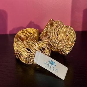 May include: A gold-colored cuff bracelet with a woven knot design. The bracelet is made of thin, intertwined strands of metal, creating a textured surface. A white tag with the "SATYA JEWELRY" logo is attached to the bracelet.