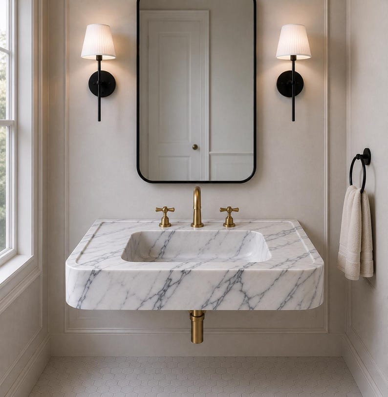 May include: A wall-mounted white marble sink with grey veining, featuring a brass faucet and two matching brass handles. Above the sink is a rectangular mirror with a black frame, flanked by two black wall sconces with white pleated shades.