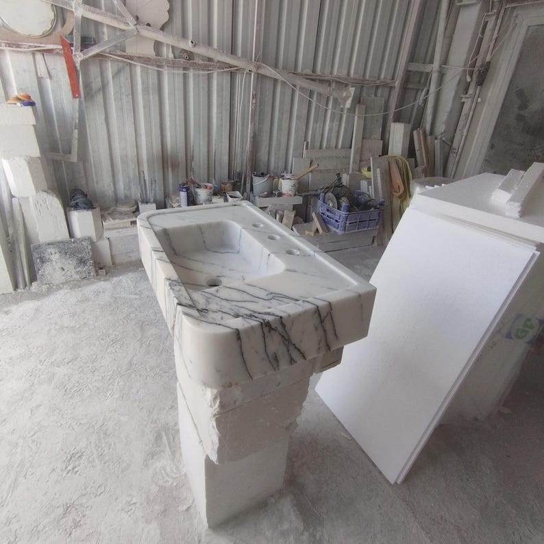May include: A rectangular pedestal sink carved from white marble with prominent dark grey veining. The basin features a smooth, polished surface with three pre-drilled faucet holes, while the base retains a rough, unfinished stone texture.