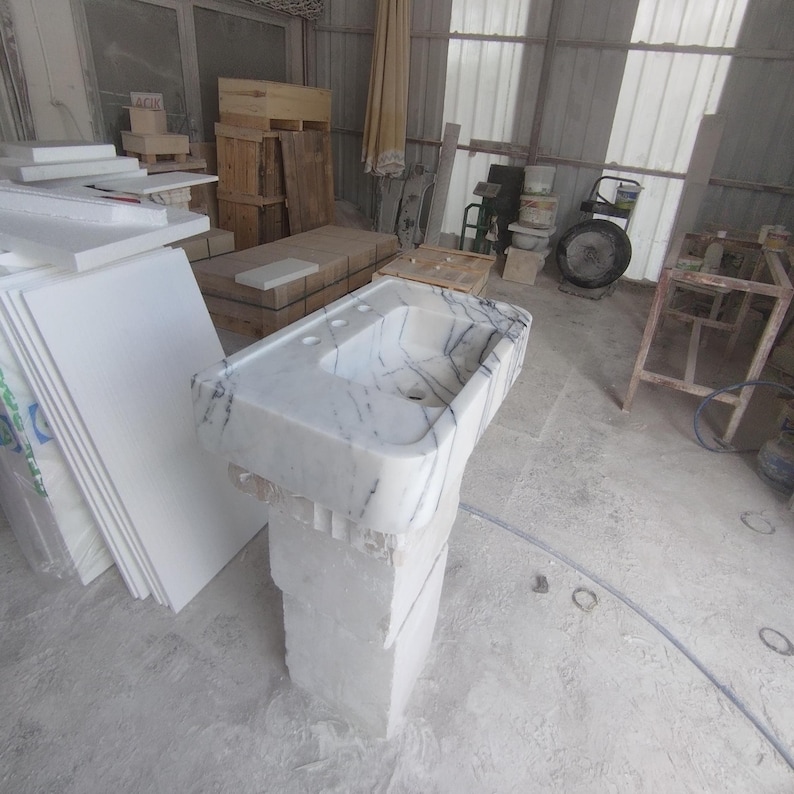 May include: A white marble pedestal sink featuring prominent dark gray veining throughout the basin and countertop. The sink has a rectangular basin with three pre-drilled faucet holes and is supported by a solid, block-like white stone base.
