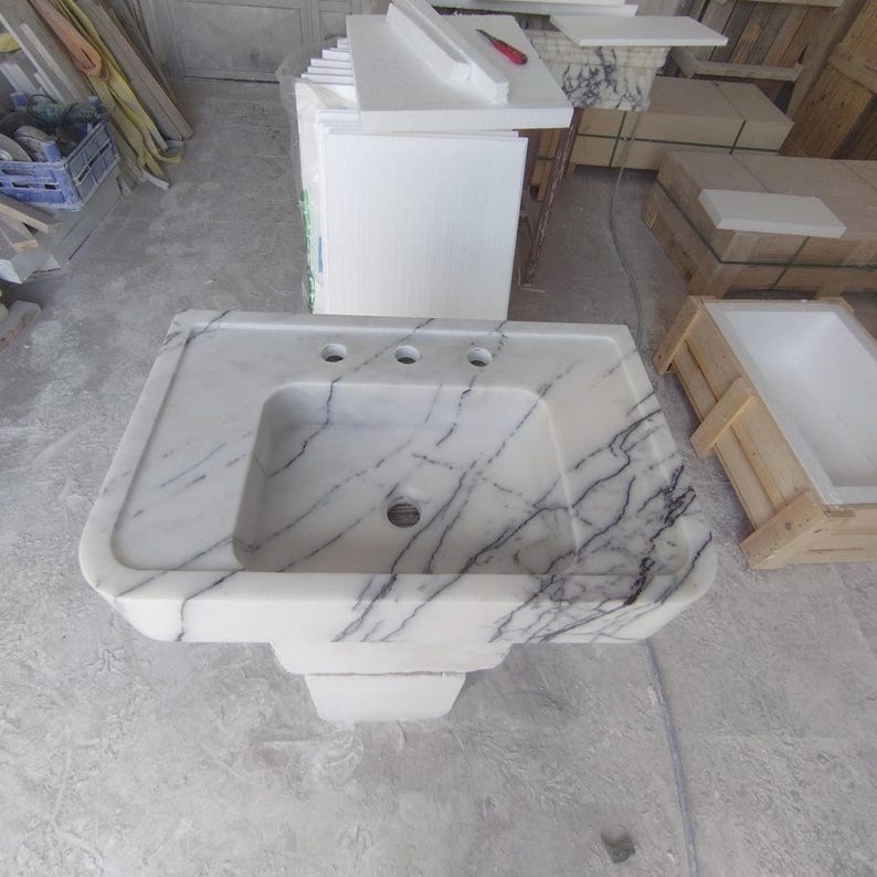 May include: A white marble pedestal sink featuring prominent black veining throughout the basin and countertop. The rectangular basin includes a central drain hole and three pre-drilled faucet holes along the back ledge of the stone structure.
