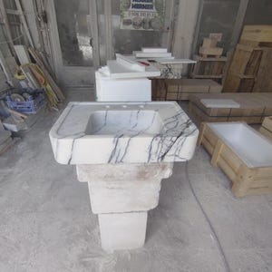 May include: A white marble pedestal sink featuring prominent dark grey veining throughout the basin and rectangular base. The sink has a wide, flat rim with three pre-drilled faucet holes and a deep, square-shaped basin, standing on a blocky stone support.