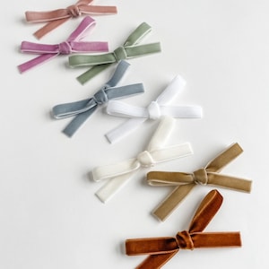 May include: Assortment of velvet hair bows in various colors, including dusty rose, sage green, white, light blue, and brown. The bows are tied in a simple knot and are perfect for adding a touch of elegance to any hairstyle.