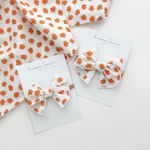 May include: Two white fabric bows with orange pumpkin patterns, displayed on white cards. The bows are part of the Kinsey Grace Collection. A matching fabric with pumpkin pattern is also visible.