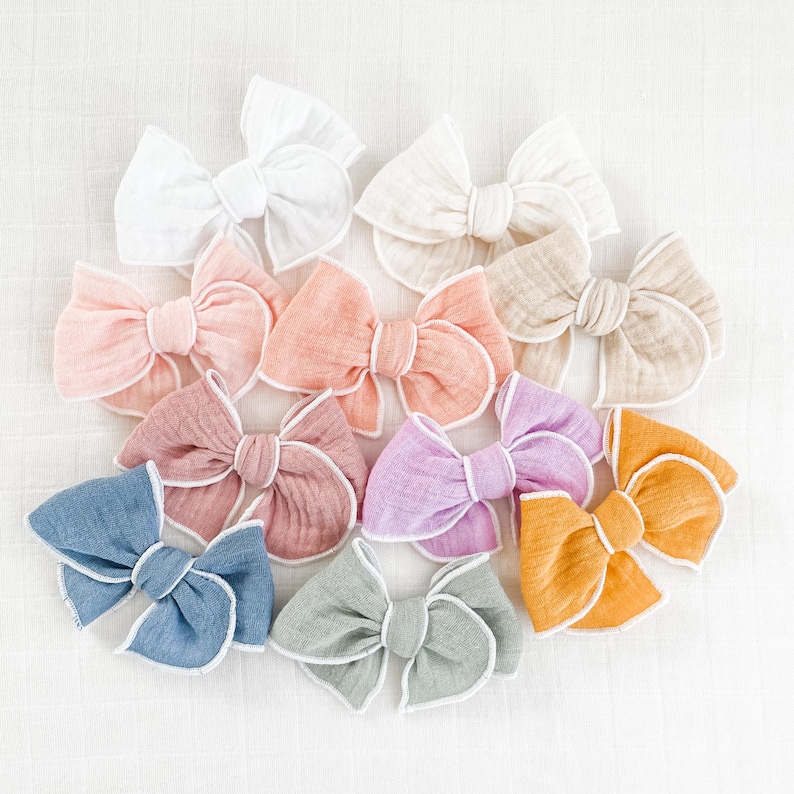 May include: A set of ten handmade fabric hair bows in various pastel colors with white trim. The bows are made of a soft, textured fabric and have a simple, elegant design.