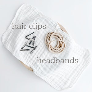 May include: A white cloth with the words "hair clips" and "headbands" printed on it. A pile of silver hair clips and a bundle of beige elastic headbands are on the cloth.