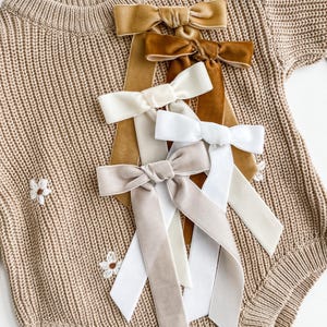 May include: A collection of five velvet hair bows in shades of tan, brown, cream, white, and light beige, each featuring long, flowing ribbon tails. The bows are arranged vertically on a beige knit garment with small white embroidered flower details.