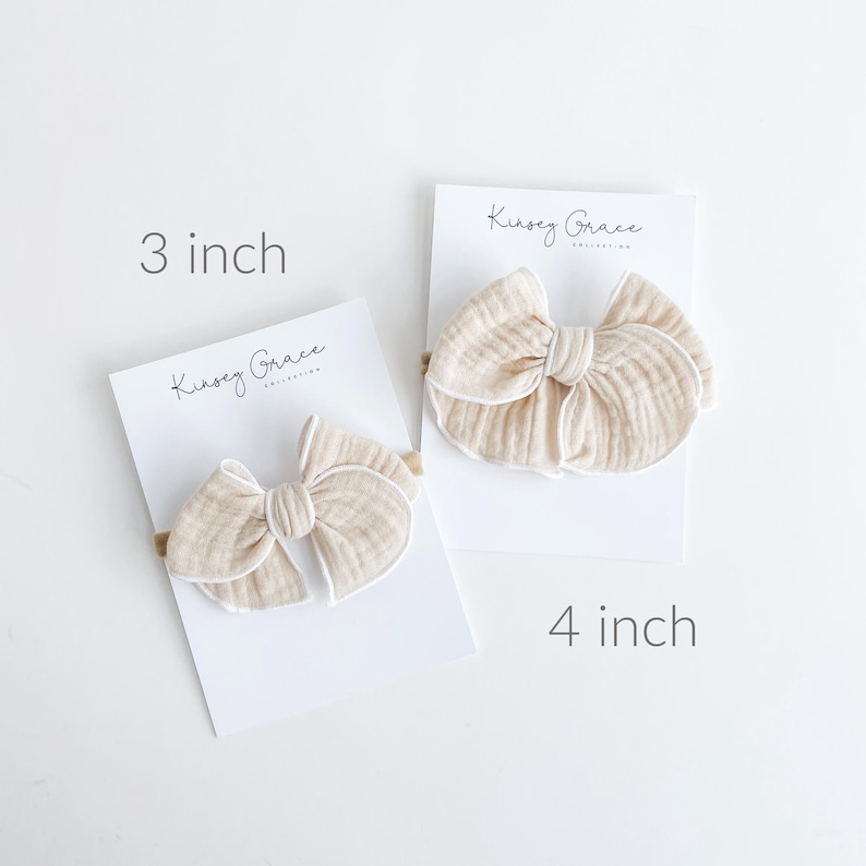 May include: Two beige fabric hair bows with white trim on white cards. The card on the left says "Kinsey Grace" and "3 inch". The card on the right says "Kinsey Grace" and "4 inch".