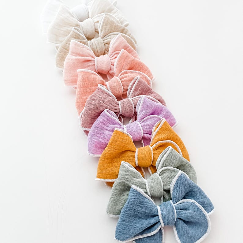 May include: A set of ten fabric hair bows in various pastel colors, each with a white trim. The bows are arranged in a line, with the lightest color at the top and the darkest color at the bottom.