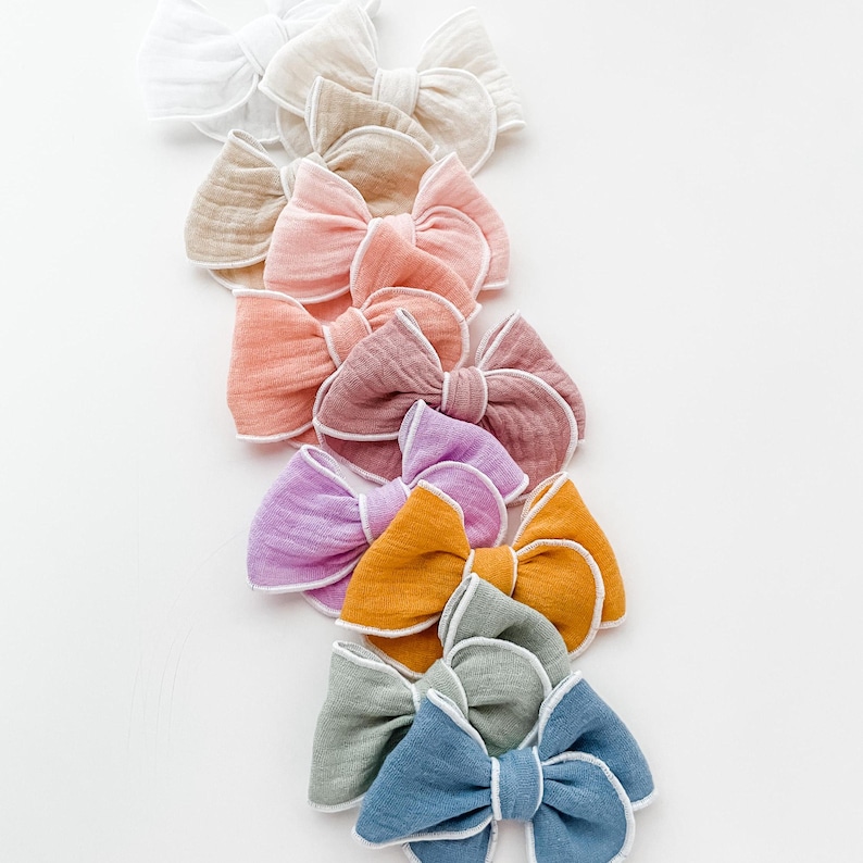 May include: A set of ten fabric hair bows in various pastel colors, each with a white trim. The bows are arranged in a vertical line, with the lightest color at the top and the darkest color at the bottom.
