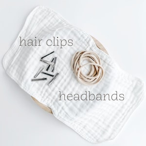 May include: A white cloth with the words "hair clips" and "headbands" printed on it. A pile of silver hair clips and a bundle of beige elastic headbands are on the cloth.