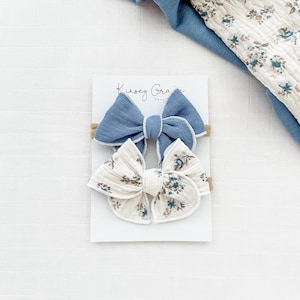 May include: Two hair bows on a white card. One bow is blue with white trim and the other is white with a floral pattern and white trim. The card has the text "Kinsey Grace" on it.