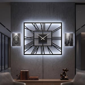 May include: A large square black wall clock with Roman numerals and a central square dial featuring thin white hands. The clock is backlit with a bright white light, creating a glowing effect against the wall, and is flanked by two small rectangular art pieces.