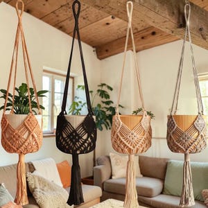 May include: Four macrame plant hangers in varying colors: peach, black, cream, and gray. Each hanger suspends a white or tan pot, some containing greenery. The hangers feature intricate woven patterns and long tassels, adding a bohemian touch.