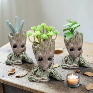 May include: Three Groot-shaped planters, each holding different types of greenery. The planters are a light brown color with detailed wood-grain texture. One holds a cactus, another has a cluster of green flowers, and the third has a small green plant. A lit candle sits nearby.
