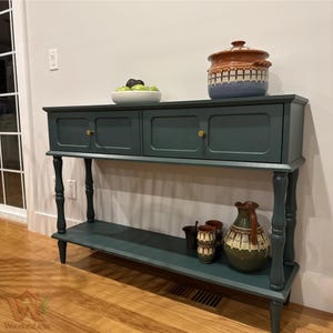 May include: A dark teal console table with two drawers, brass knobs, and a lower shelf. On top sits a white bowl with green fruit and a lidded ceramic pot. The lower shelf holds a ceramic pitcher and three small matching cups.