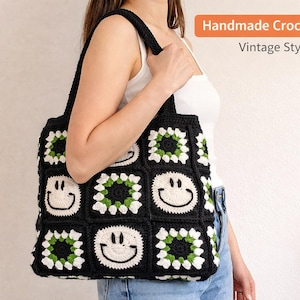 May include: Handmade black crochet tote bag with white smiley faces and green accents. The bag has a vintage style and features a square pattern. The bag has a black strap and is held by a person.