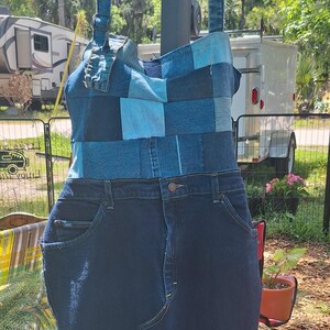 May include: A denim dress with a patchwork bodice in shades of blue and a dark blue denim skirt. The dress has adjustable straps and is hanging on a black hanger. The dress is made from recycled denim.