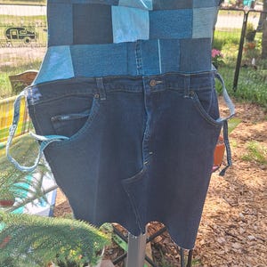 May include: A denim apron crafted from recycled jeans. The top section is patchwork, with different shades of blue denim. The bottom half keeps the original jean pockets and button fastening. Adjustable straps complete the design.