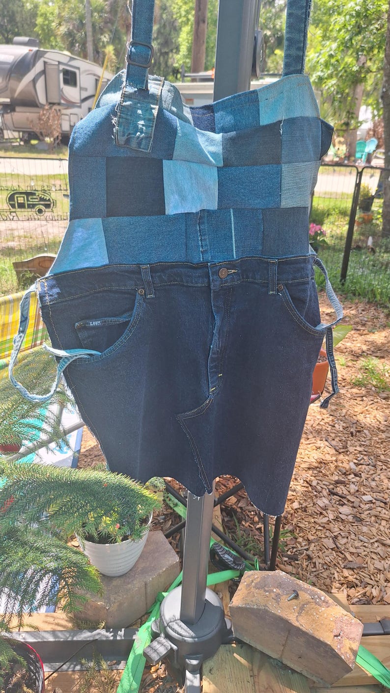 May include: A denim apron crafted from recycled jeans. The top section is patchwork, with different shades of blue denim. The bottom half keeps the original jean pockets and button fastening. Adjustable straps complete the design.