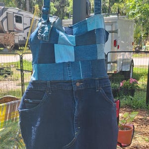 May include: A denim dress with a patchwork bodice in shades of blue and a dark blue denim skirt. The dress has adjustable straps and is hanging on a black hanger. The dress is made from recycled denim.