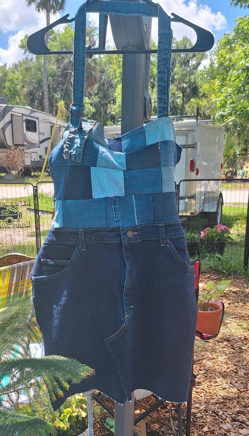 May include: A denim dress with a patchwork bodice in shades of blue and a dark blue denim skirt. The dress has adjustable straps and is hanging on a black hanger. The dress is made from recycled denim.