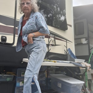 May include: Light blue denim jacket with floral embroidery and matching jeans. The jeans feature a patchwork design on the side, with a flared leg. The person is wearing a red top underneath the jacket.