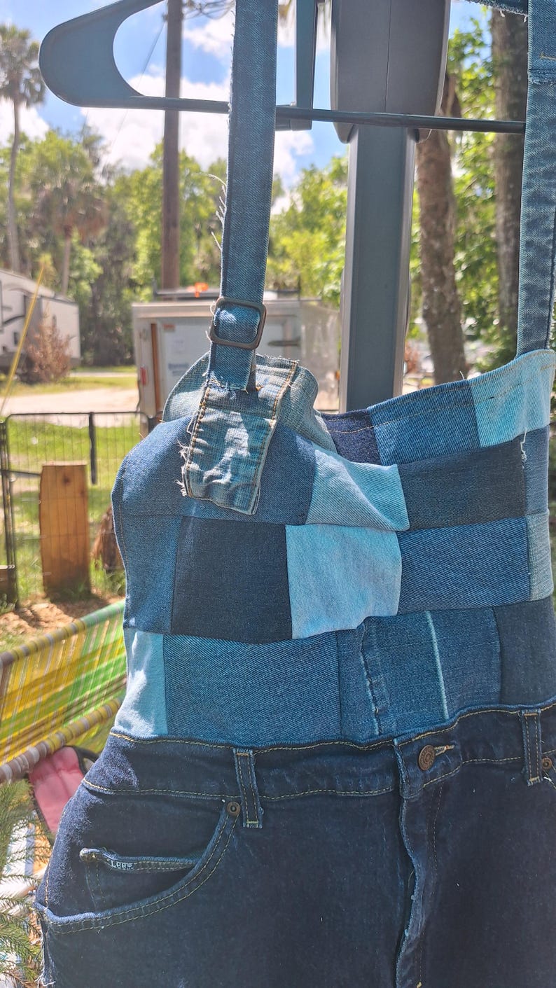 May include: A denim shoulder bag, crafted from patches of blue denim in various shades. The bag features a strap with a buckle and incorporates the waistband and pockets of a pair of jeans at the base. The bag is a unique, upcycled design.