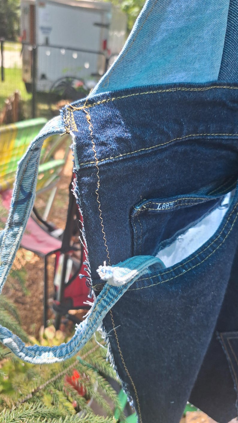 May include: Close-up of a denim bag with a patchwork design. The bag features various shades of blue denim, with visible stitching in yellow thread. The bag has a strap made from a lighter blue denim material. The Lee logo is visible on the pocket.