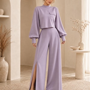 Puede incluir: Un conjunto de dos piezas color lavanda que consta de un top corto de manga larga con cuello alto y un bolsillo en el pecho, junto con pantalones de talle alto y pierna ancha con una abertura vertical en la pierna derecha y pu&ntilde;os abotonados.