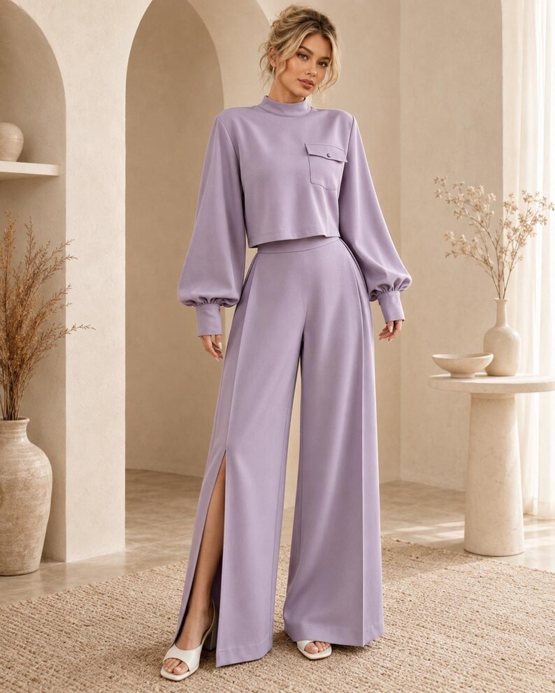 Puede incluir: Un conjunto de dos piezas color lavanda que consta de un top corto de manga larga con cuello alto y un bolsillo en el pecho, junto con pantalones de talle alto y pierna ancha con una abertura vertical en la pierna derecha y pu&ntilde;os abotonados.